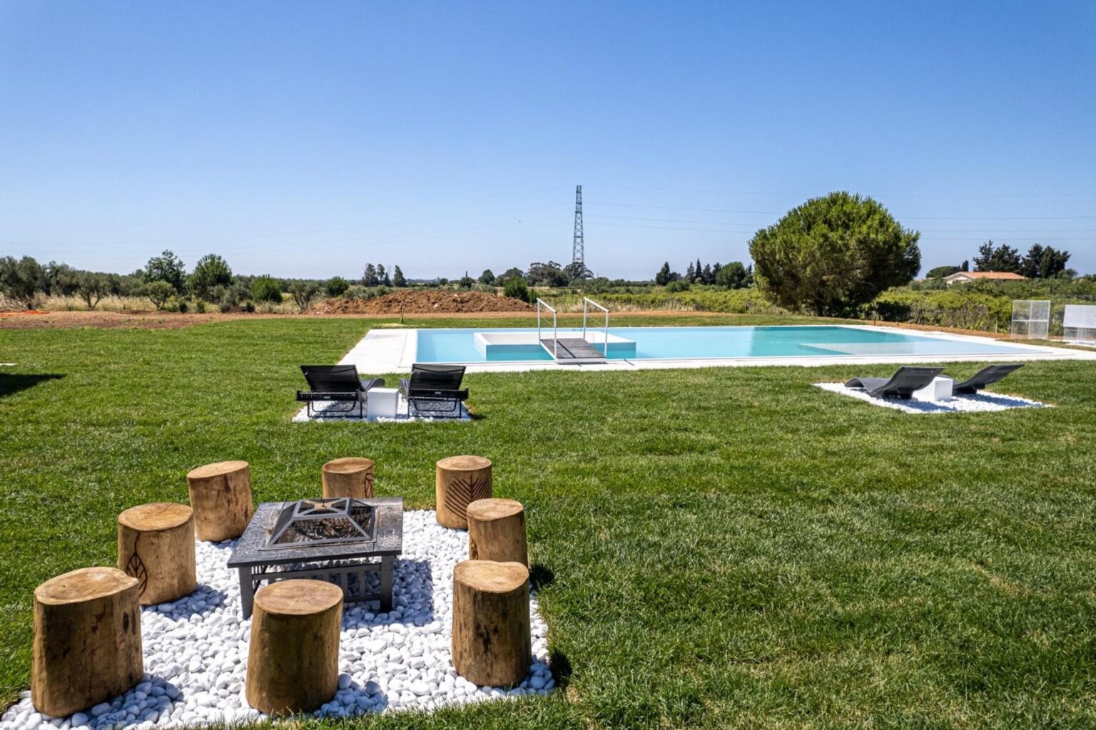 Outdoor pool area featuring a well-maintained lawn, lounge chairs, and a cozy fire pit surrounded by wooden stools, ideal for relaxation and gatherings. | Only on SopranoVillas
