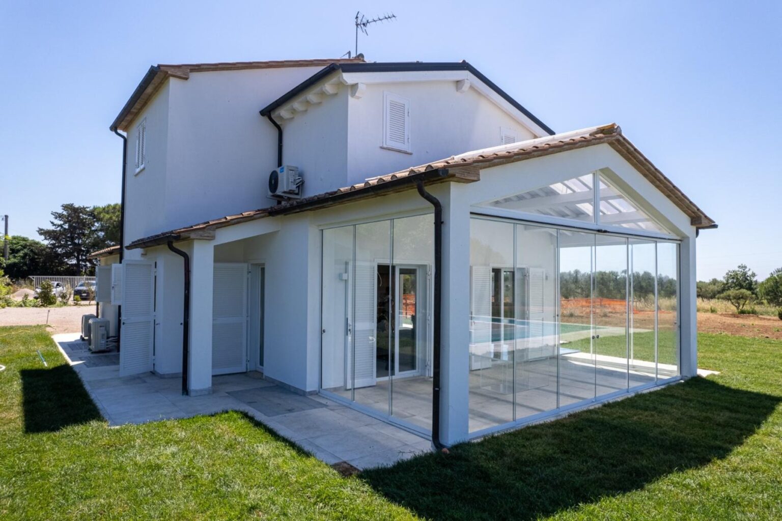 Modern two-story house with a glass-enclosed porch, surrounded by green grass and a clear blue sky. The exterior features a white facade, tiled roof, and air conditioning unit, showcasing contemporary architecture in a serene rural setting. | Only on SopranoVillas