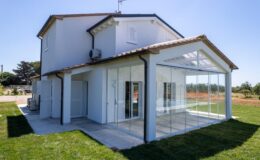 Modern two-story house with a glass-enclosed porch, surrounded by green grass and a clear blue sky. The exterior features a white facade, tiled roof, and air conditioning unit, showcasing contemporary architecture in a serene rural setting.