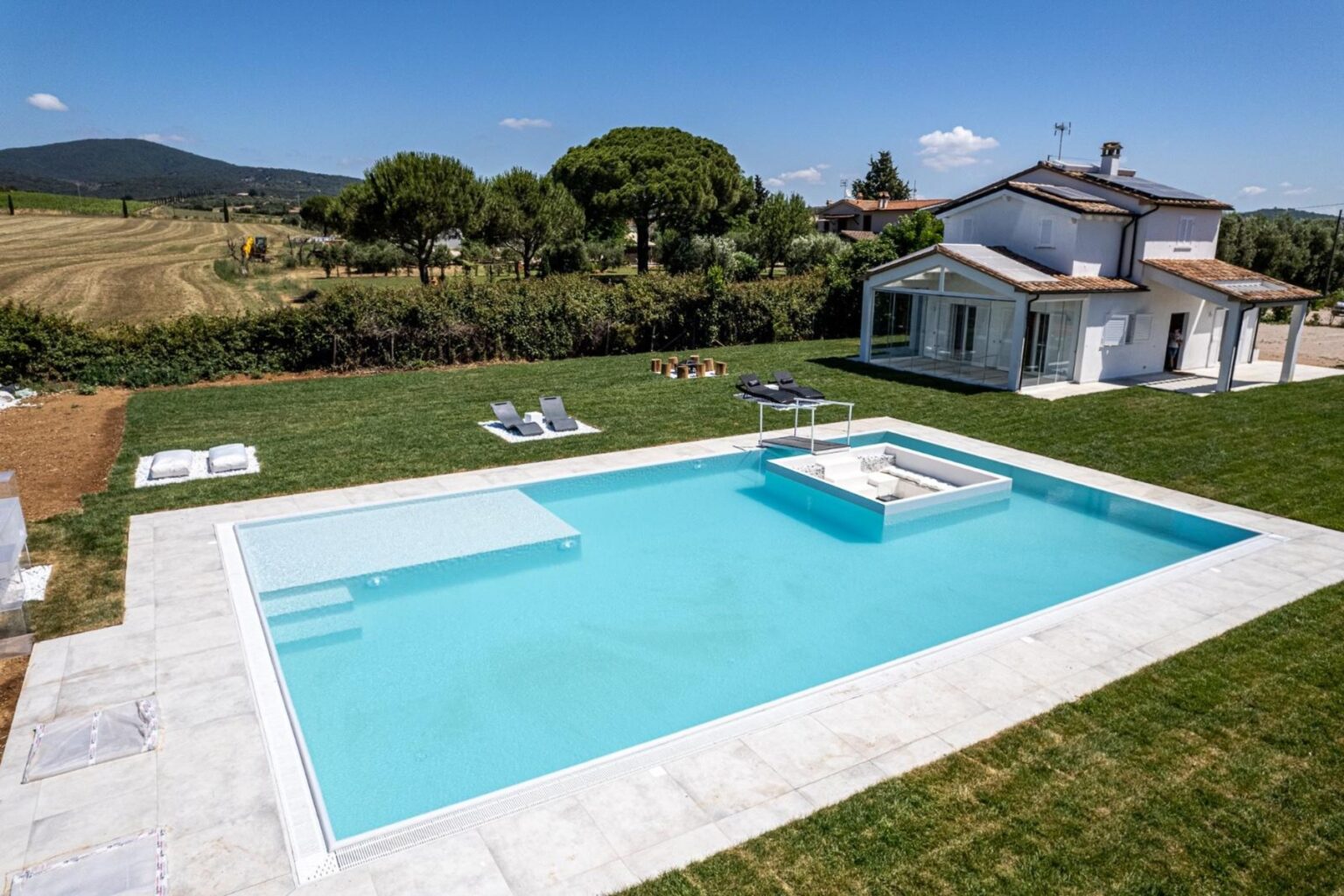 Modern villa with a spacious outdoor pool surrounded by lush greenery and a scenic landscape, featuring lounge chairs for relaxation and a sunny blue sky. | Only on SopranoVillas