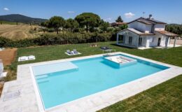 Modern villa with a spacious outdoor pool surrounded by lush greenery and a scenic landscape, featuring lounge chairs for relaxation and a sunny blue sky.