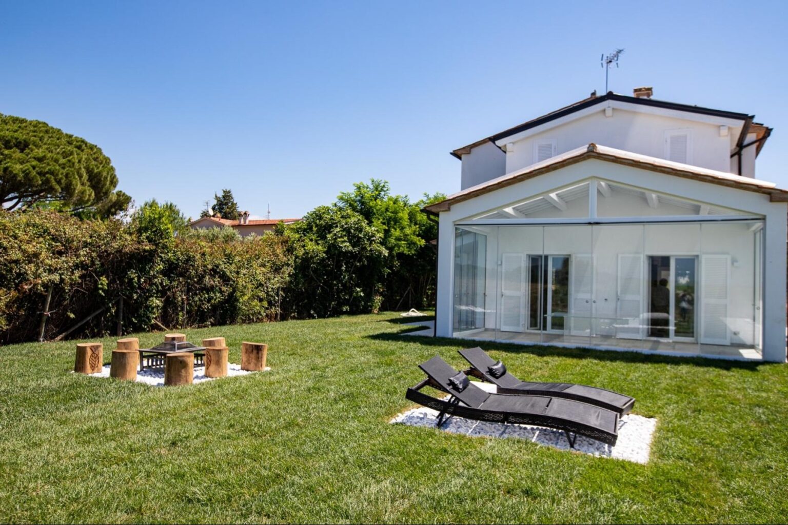 Modern house with a glass extension set in a lush green garden, featuring lounge chairs and a fire pit area, perfect for outdoor relaxation and entertaining. | Only on SopranoVillas