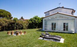 Modern house with a glass extension set in a lush green garden, featuring lounge chairs and a fire pit area, perfect for outdoor relaxation and entertaining.