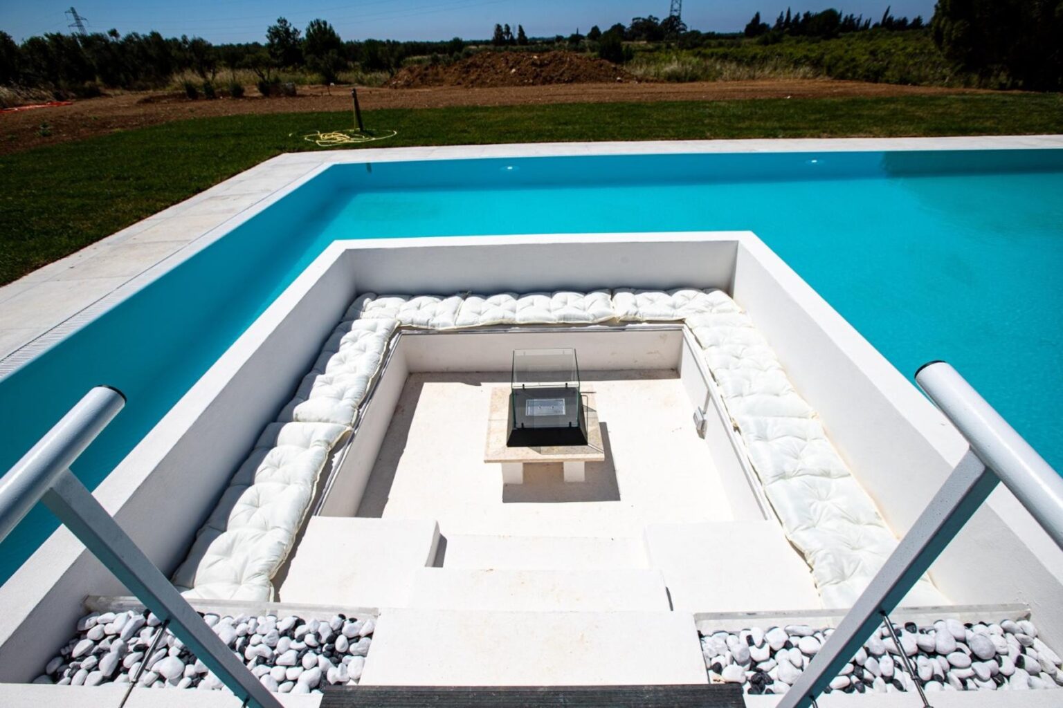 Modern outdoor pool design featuring a white stone fire pit surrounded by a stylish seating area, with clear blue water and lush green landscape in the background. Ideal for relaxation and entertainment. | Only on SopranoVillas