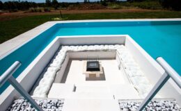 Modern outdoor pool design featuring a white stone fire pit surrounded by a stylish seating area, with clear blue water and lush green landscape in the background. Ideal for relaxation and entertainment.