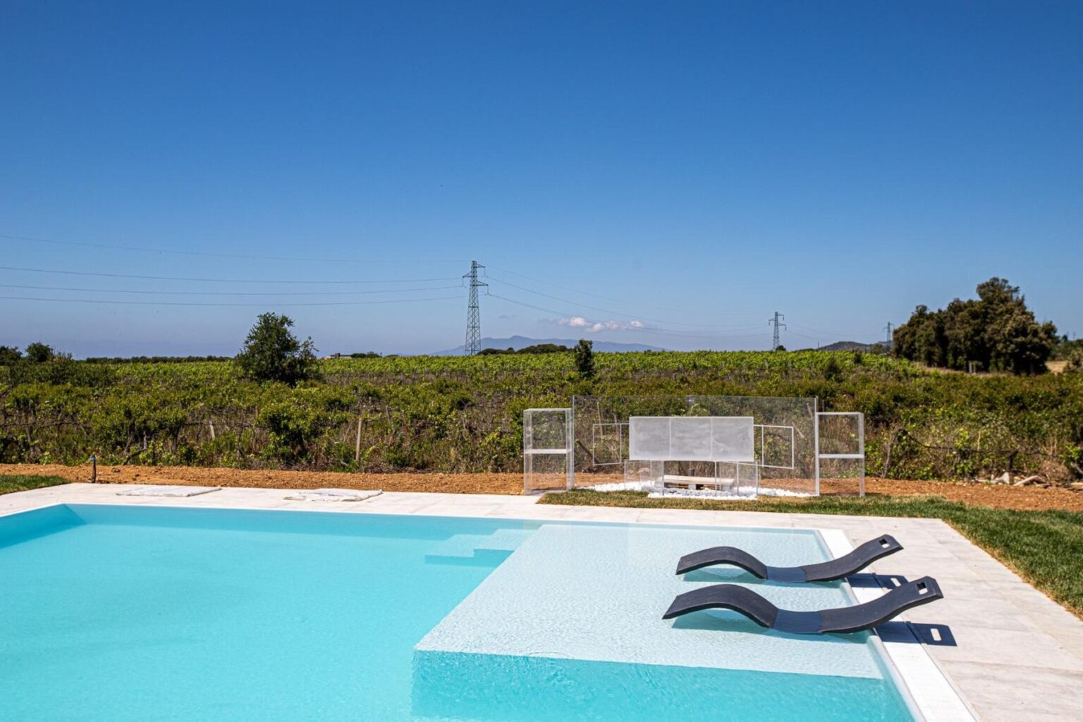Outdoor swimming pool with two lounge chairs, surrounded by a scenic landscape of greenery and distant hills under a clear blue sky. Ideal for relaxation and leisure activities. | Only on SopranoVillas