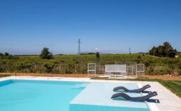 Outdoor swimming pool with two lounge chairs, surrounded by a scenic landscape of greenery and distant hills under a clear blue sky. Ideal for relaxation and leisure activities.