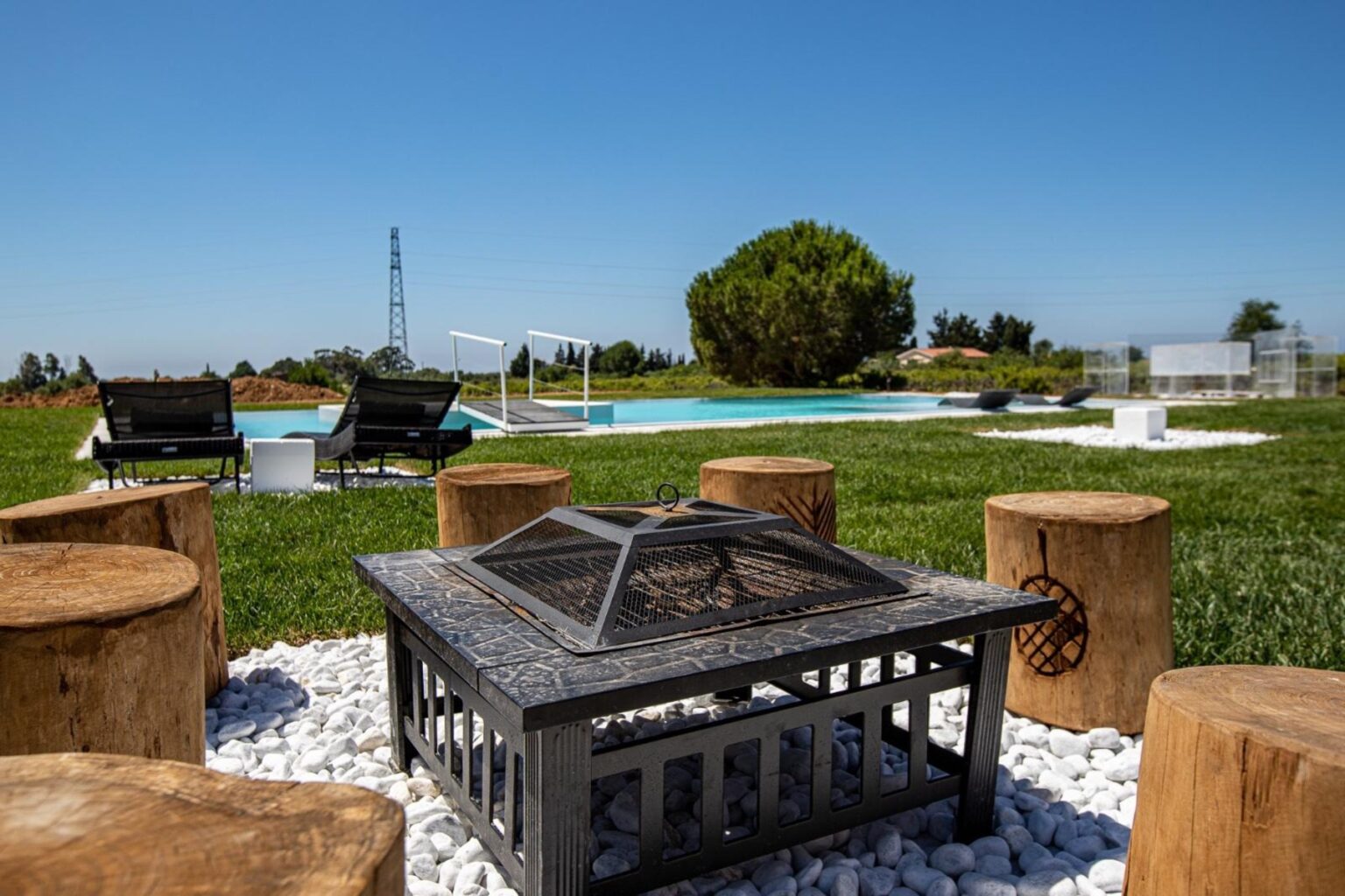 Cozy outdoor seating area with a fire pit surrounded by wooden stools, overlooking a swimming pool and lush green landscape under a clear blue sky. | Only on SopranoVillas