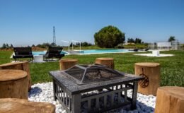 Cozy outdoor seating area with a fire pit surrounded by wooden stools, overlooking a swimming pool and lush green landscape under a clear blue sky.