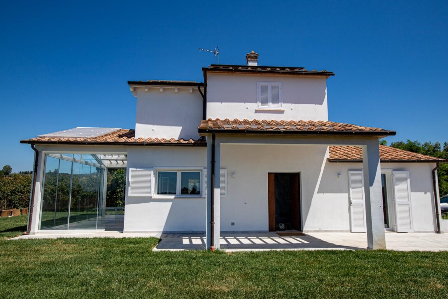 Modern two-story house with a tiled roof, featuring a glass-enclosed porch and a spacious lawn under a clear blue sky. | Only on SopranoVillas