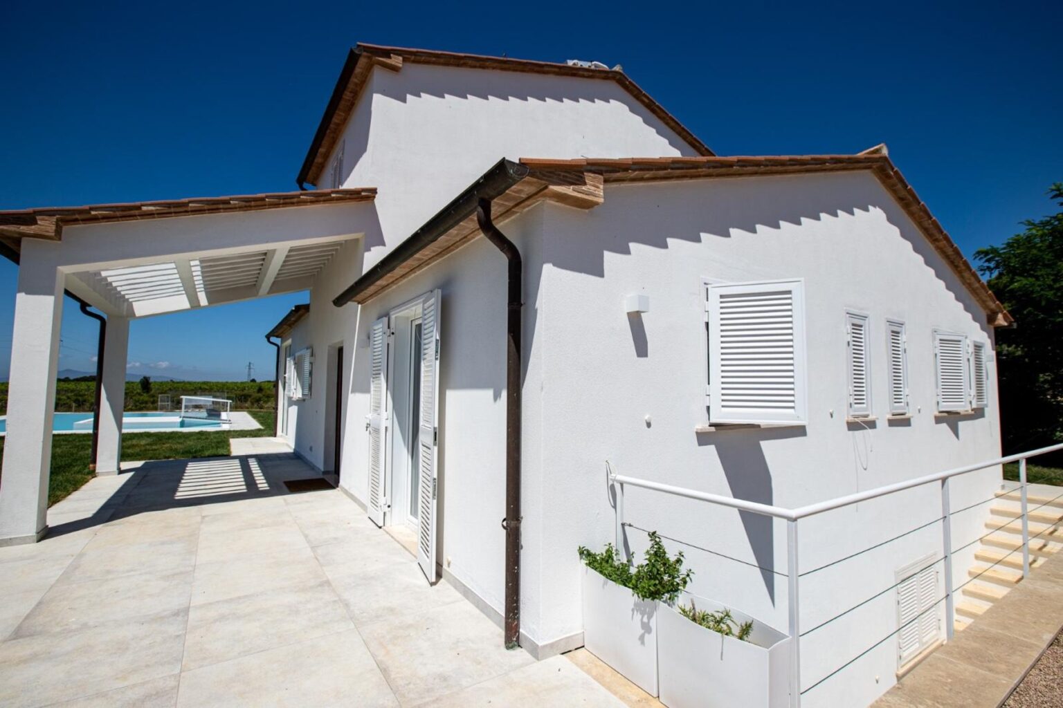 Modern white house exterior with a sloped roof, featuring large windows and a spacious patio area, surrounded by green landscaping under a clear blue sky. | Only on SopranoVillas