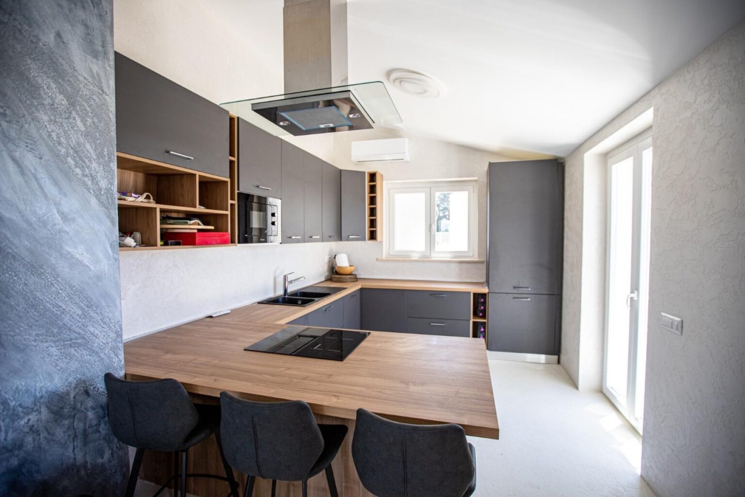 Modern kitchen featuring sleek gray cabinets, a wooden island with a built-in cooktop, and large windows for natural light. | Only on SopranoVillas