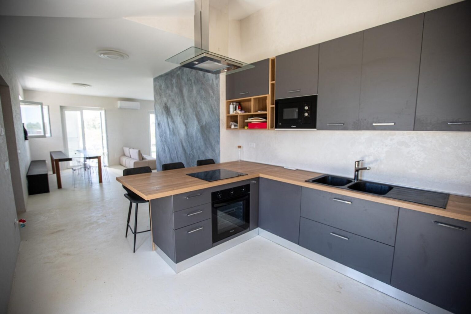 Modern kitchen interior featuring sleek gray cabinetry, a wooden countertop, and stainless steel appliances. The open layout connects to a bright dining area with natural light streaming in from large windows. | Only on SopranoVillas