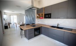 Modern kitchen interior featuring sleek gray cabinetry, a wooden countertop, and stainless steel appliances. The open layout connects to a bright dining area with natural light streaming in from large windows.