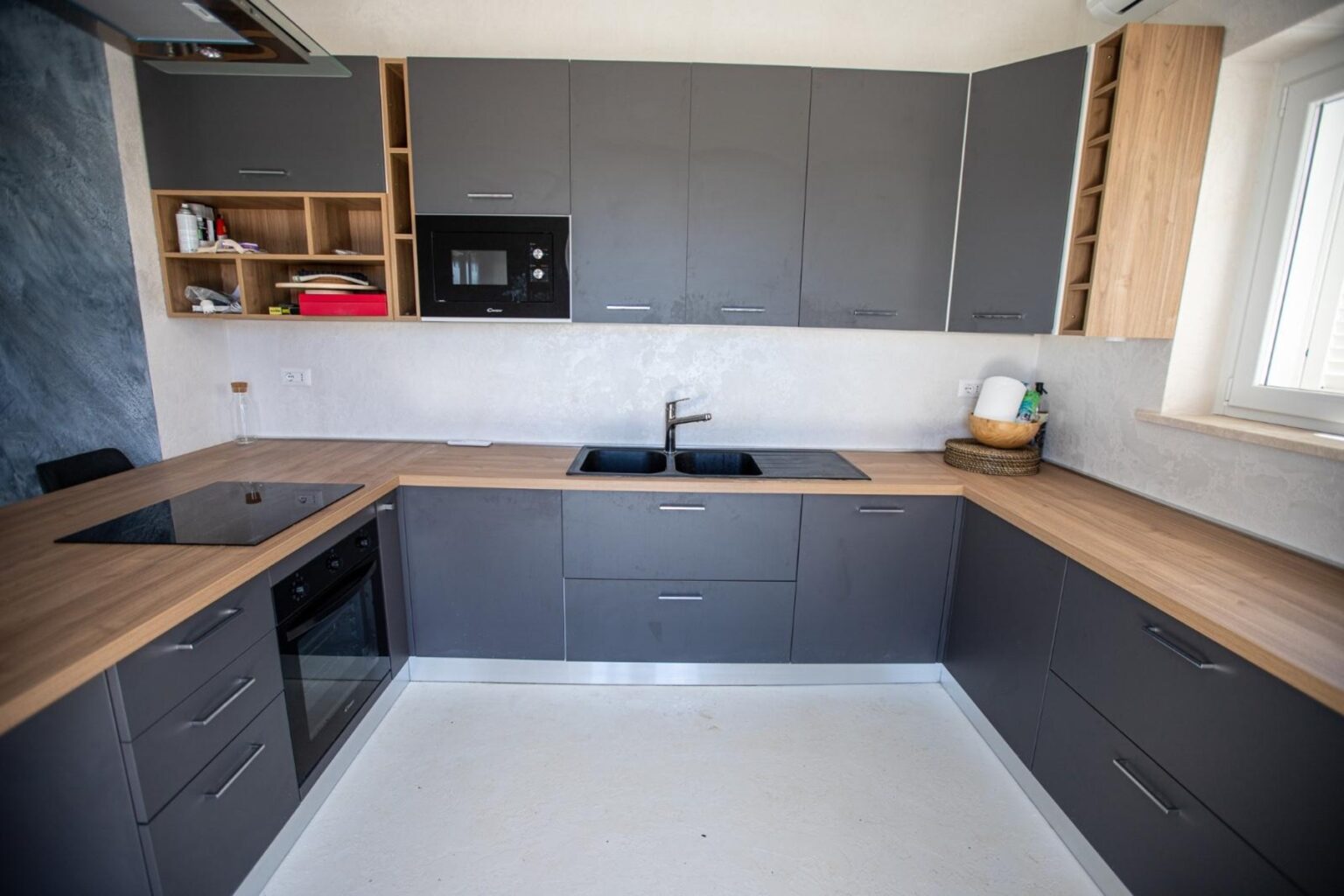 Modern kitchen featuring gray cabinets, a black sink, and wooden countertops. The design includes built-in appliances and ample storage space, creating a stylish and functional cooking area. Natural light illuminates the space through a window. | Only on SopranoVillas