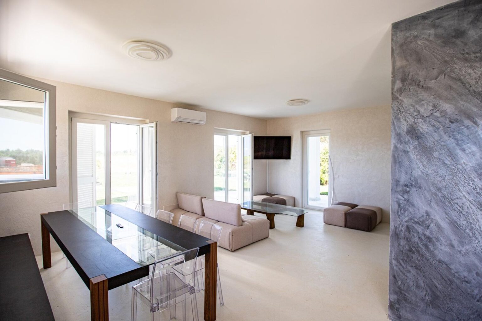 Modern living room with large windows, neutral-colored walls, a comfortable sofa, and a sleek glass table, creating a bright and airy atmosphere. | Only on SopranoVillas