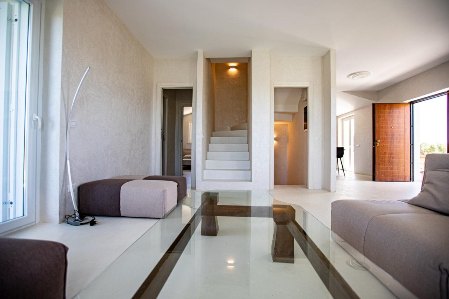 Modern living room interior featuring a glass coffee table, minimalist furniture, and a staircase in the background, with natural light streaming through large windows. | Only on SopranoVillas