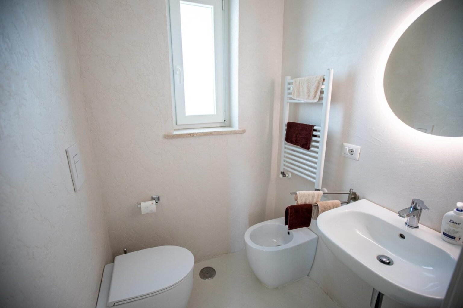 Modern bathroom interior featuring a toilet, bidet, and a sleek sink. Natural light enters through a window, with a towel warmer and neatly arranged towels enhancing the space's functionality and style. Soft, neutral colors create a calming atmosphere. | Only on SopranoVillas
