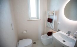 Modern bathroom interior featuring a toilet, bidet, and a sleek sink. Natural light enters through a window, with a towel warmer and neatly arranged towels enhancing the space's functionality and style. Soft, neutral colors create a calming atmosphere.
