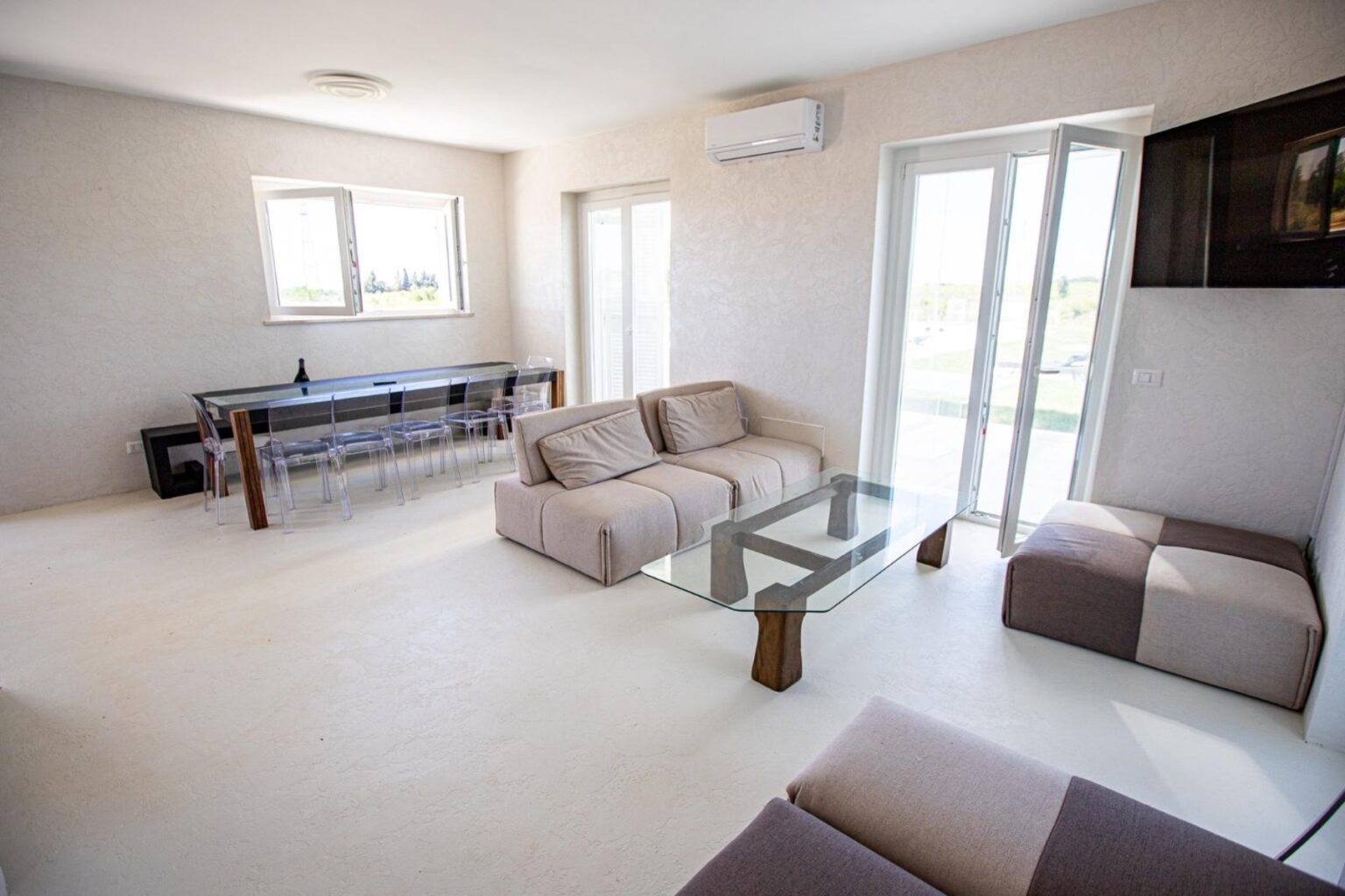 Modern living room with minimalistic design featuring a light color palette, comfortable sofas, a glass coffee table, and large windows allowing natural light. The space includes a dining area with a sleek table and chairs, creating a bright and airy atmosphere. | Only on SopranoVillas