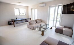 Modern living room with minimalistic design featuring a light color palette, comfortable sofas, a glass coffee table, and large windows allowing natural light. The space includes a dining area with a sleek table and chairs, creating a bright and airy atmosphere.
