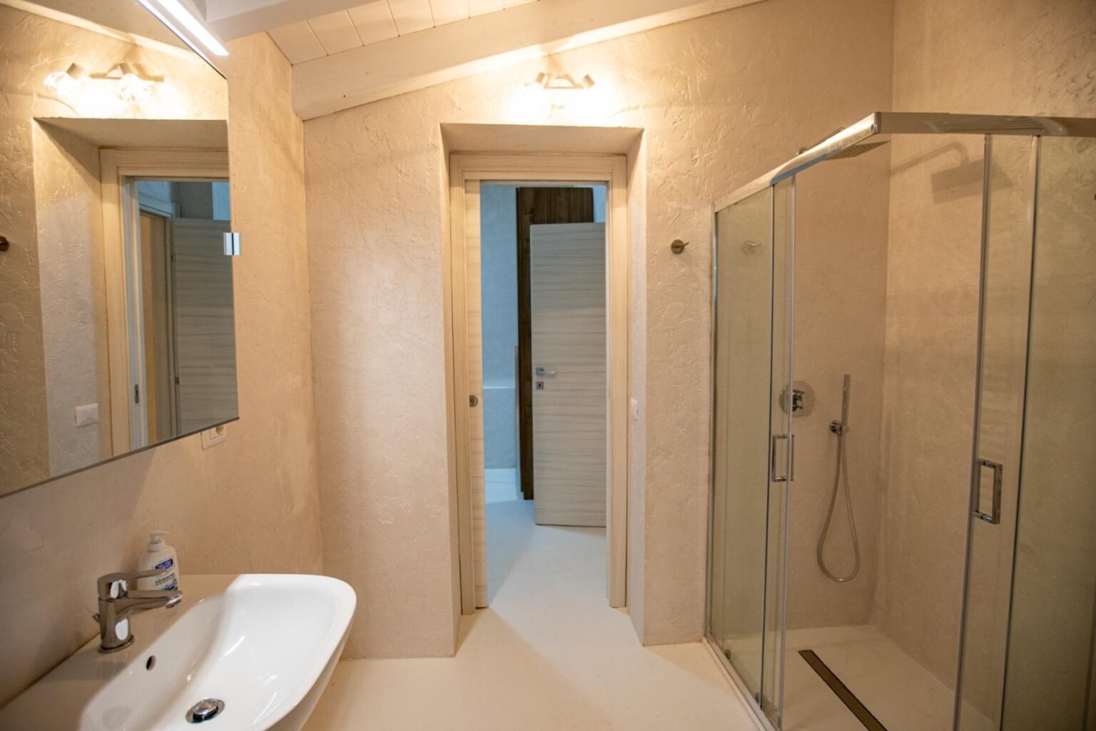 Modern bathroom featuring a sleek design with a glass shower enclosure, a white sink with a contemporary faucet, and textured beige walls. The space is well-lit with overhead lighting and includes a mirror above the sink. | Only on SopranoVillas