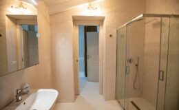 Modern bathroom featuring a sleek design with a glass shower enclosure, a white sink with a contemporary faucet, and textured beige walls. The space is well-lit with overhead lighting and includes a mirror above the sink.