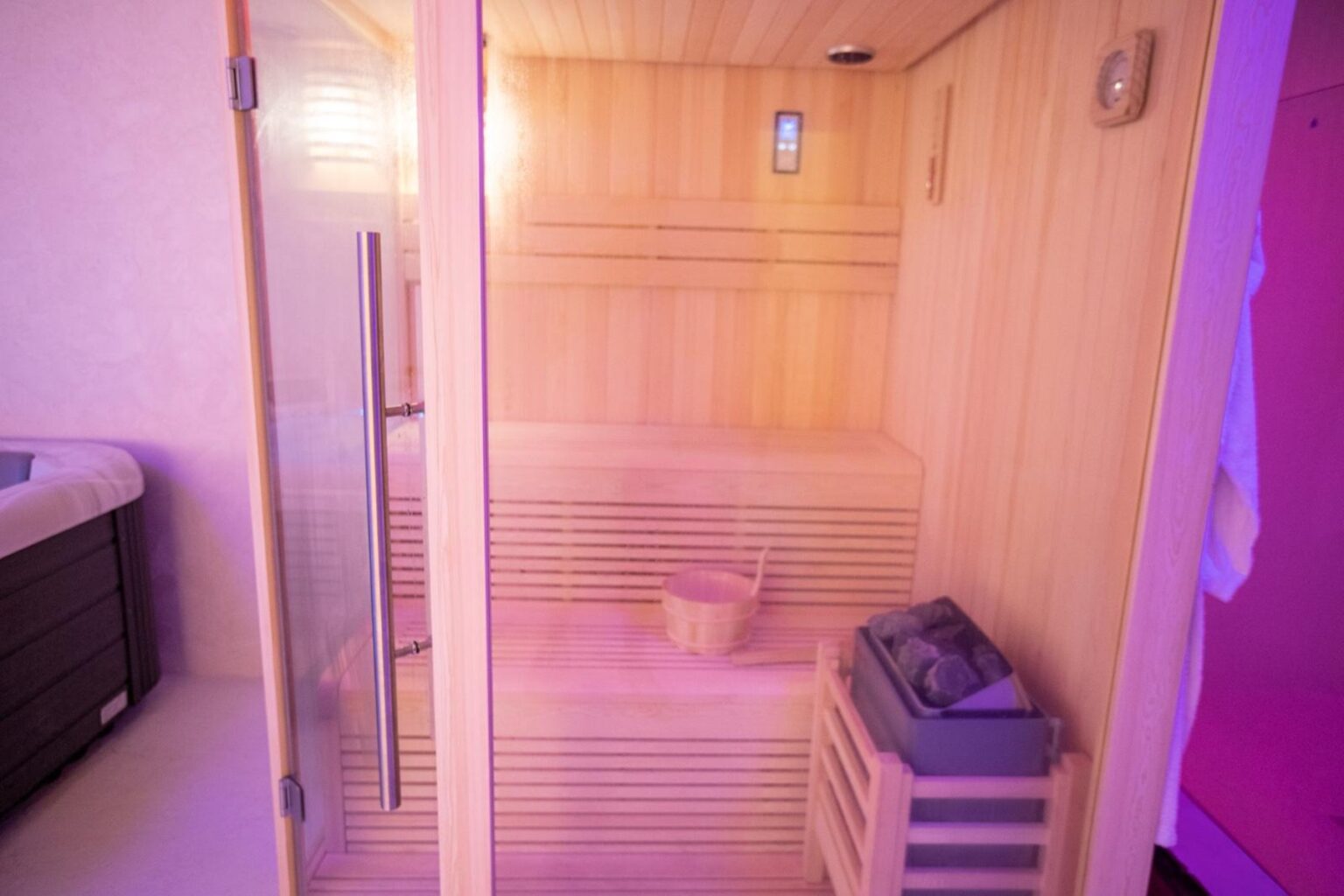 Interior of a modern sauna featuring wooden benches, a bucket for water, and a heater, illuminated with soft purple lighting. | Only on SopranoVillas