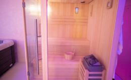 Interior of a modern sauna featuring wooden benches, a bucket for water, and a heater, illuminated with soft purple lighting.