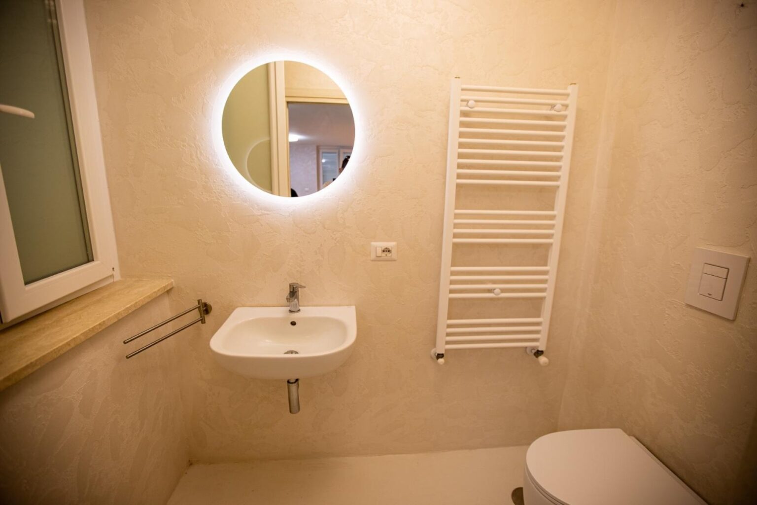 Modern bathroom interior featuring a round illuminated mirror, a compact sink, a towel radiator, and a clean, minimalist design. The light-colored walls enhance the spacious feel of the room. | Only on SopranoVillas