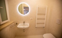 Modern bathroom interior featuring a round illuminated mirror, a compact sink, a towel radiator, and a clean, minimalist design. The light-colored walls enhance the spacious feel of the room.