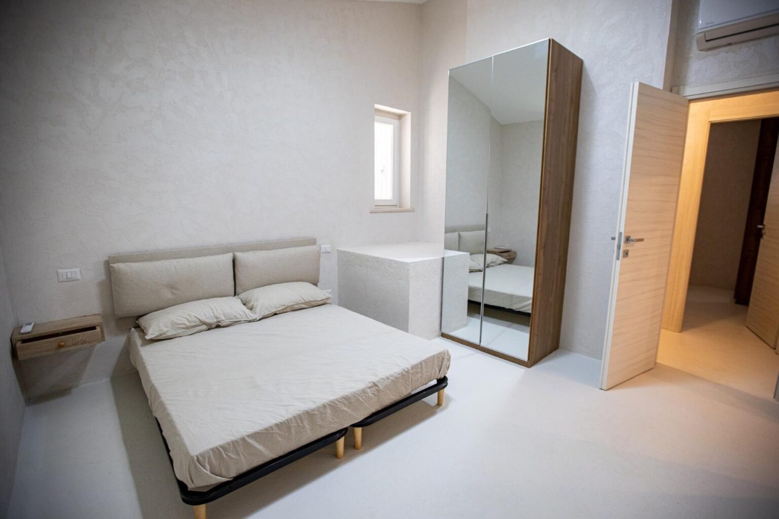 Modern bedroom interior featuring a minimalist design, a queen-sized bed with neutral bedding, a sleek mirrored wardrobe, and a light-colored wall. Natural light enters through a small window, creating a bright and airy atmosphere. | Only on SopranoVillas