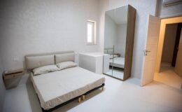 Modern bedroom interior featuring a minimalist design, a queen-sized bed with neutral bedding, a sleek mirrored wardrobe, and a light-colored wall. Natural light enters through a small window, creating a bright and airy atmosphere.