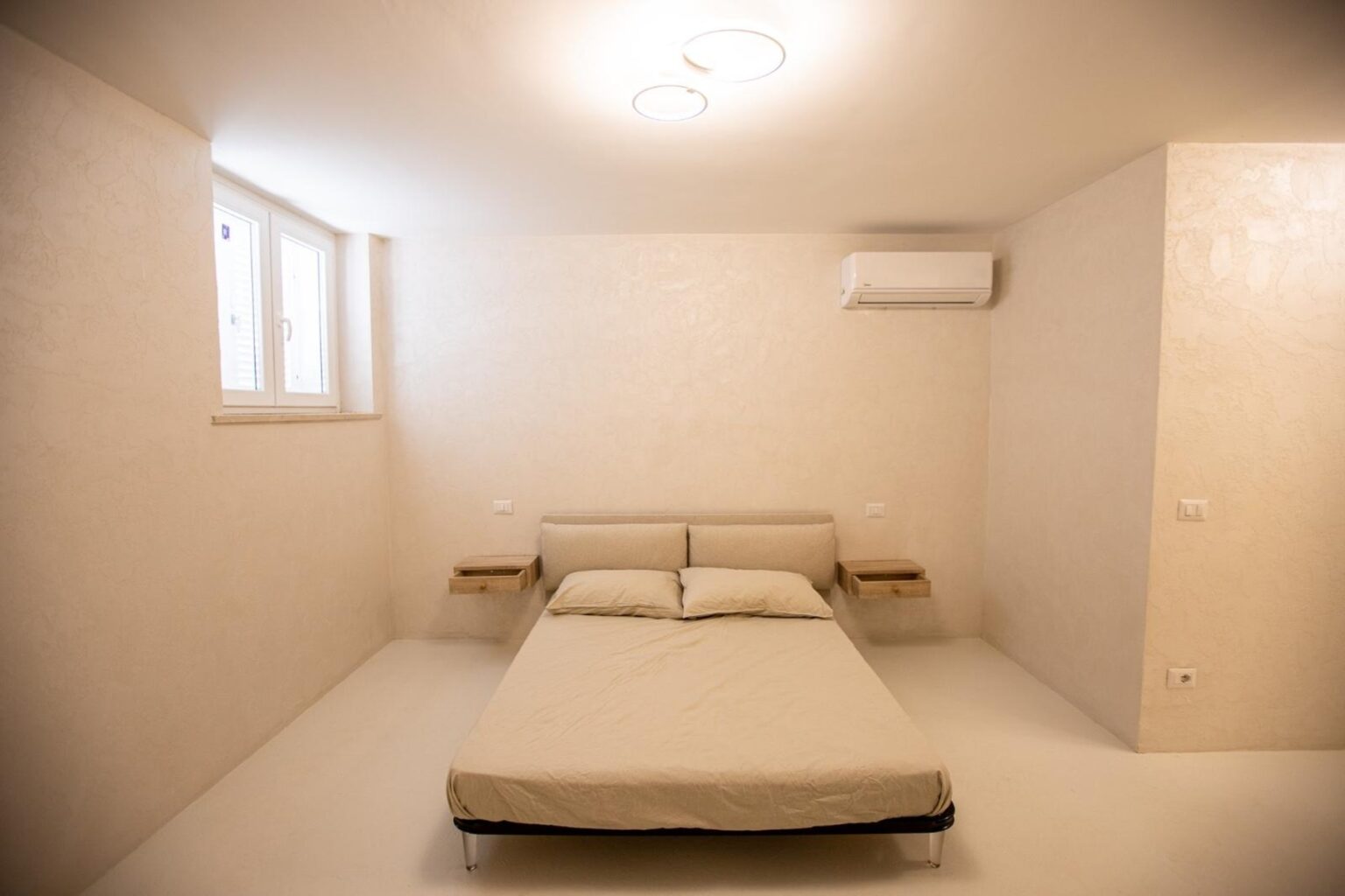 Minimalist bedroom design featuring a double bed with soft linens, wall-mounted bedside tables, and an air conditioning unit in a light-colored, modern space with natural light from windows. | Only on SopranoVillas