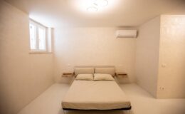Minimalist bedroom design featuring a double bed with soft linens, wall-mounted bedside tables, and an air conditioning unit in a light-colored, modern space with natural light from windows.