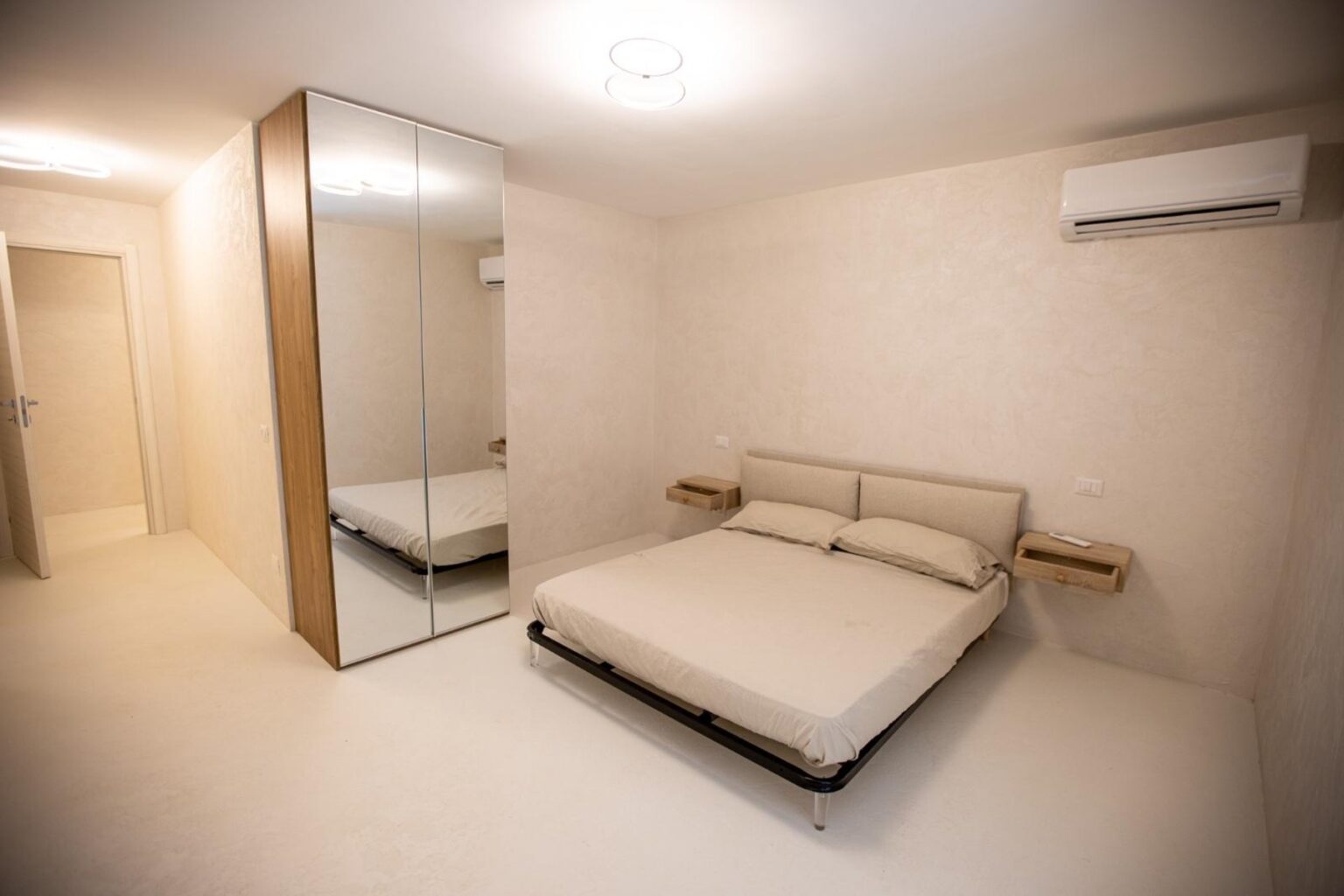 Modern bedroom interior featuring a minimalist design, a double bed with neutral bedding, a large mirrored wardrobe, and an air conditioning unit, all set against soft beige walls and a light-colored floor. | Only on SopranoVillas