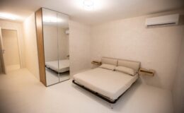 Modern bedroom interior featuring a minimalist design, a double bed with neutral bedding, a large mirrored wardrobe, and an air conditioning unit, all set against soft beige walls and a light-colored floor.