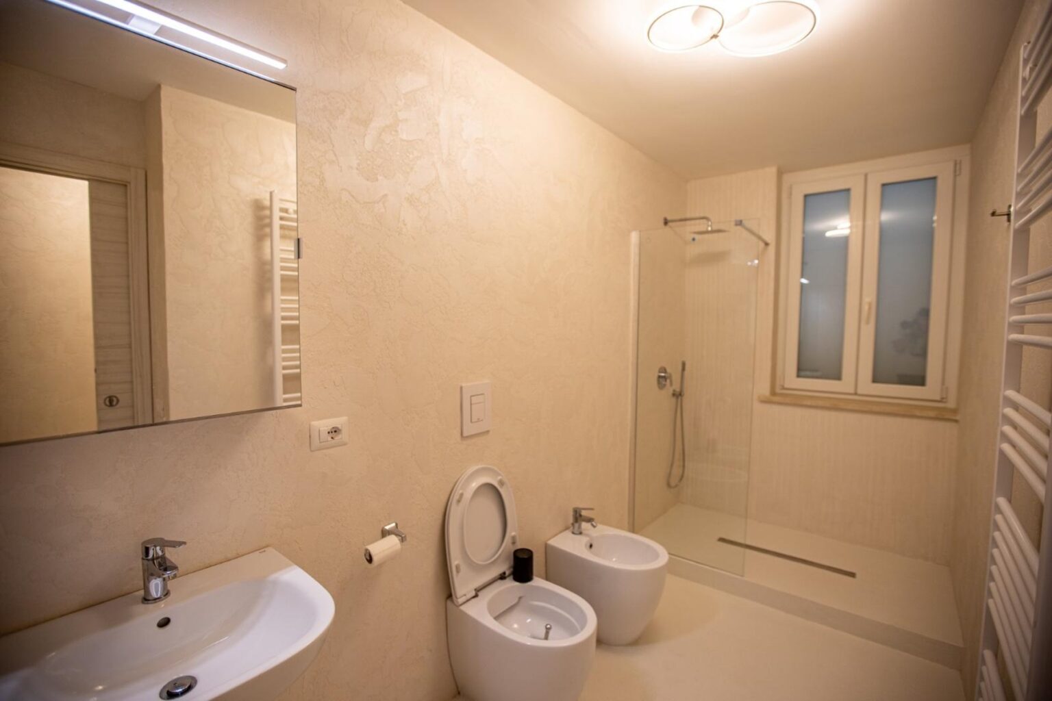 Modern bathroom featuring a shower, bidet, and sleek vanity with a mirror, illuminated by natural light from a nearby window. The neutral color palette enhances the spacious feel. | Only on SopranoVillas
