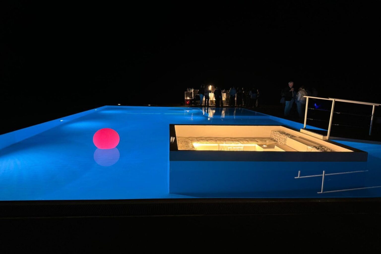 Modern swimming pool at night with illuminated blue water, featuring a bright red floating ball and a spa area. | Only on SopranoVillas