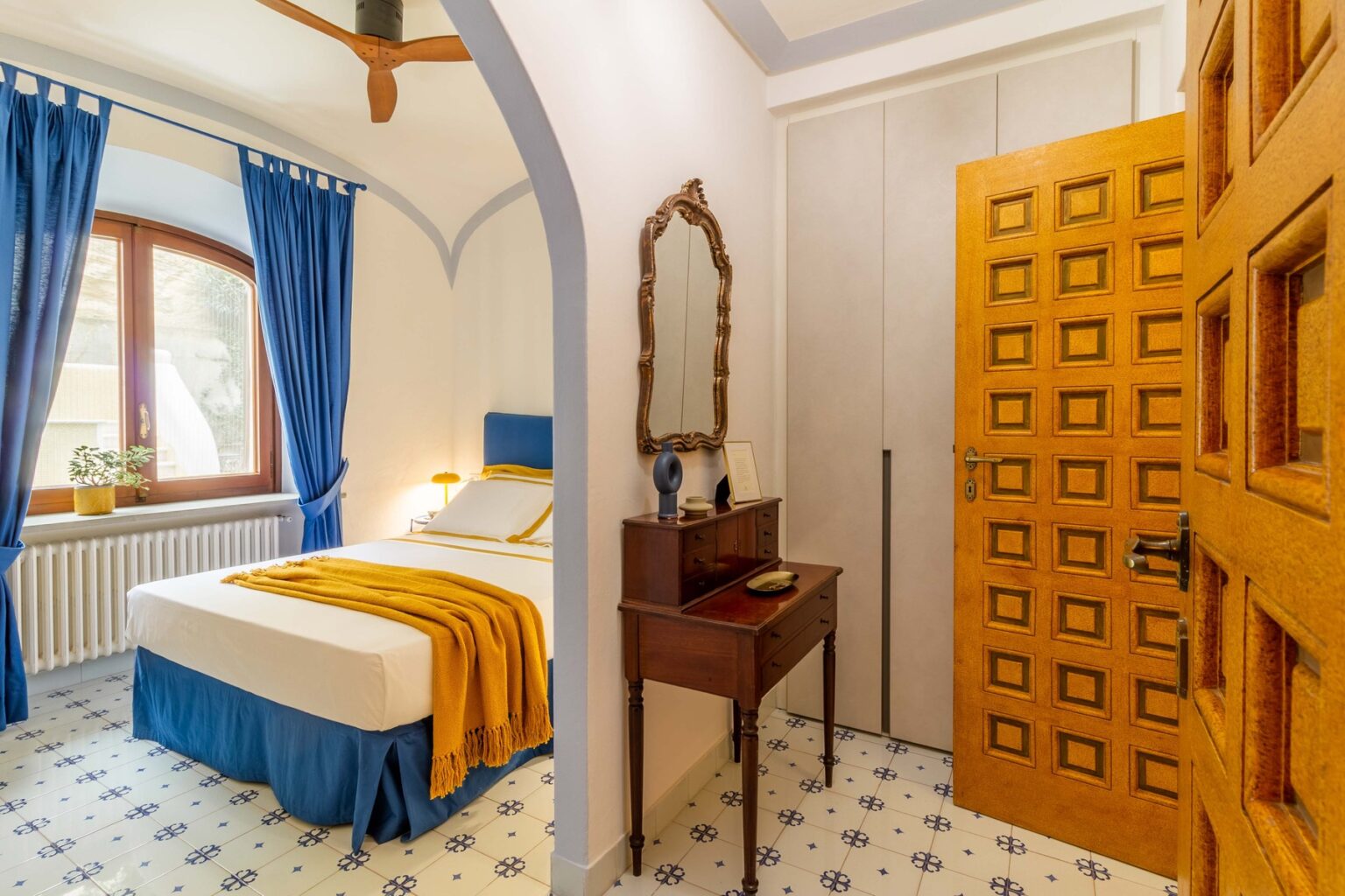 Cozy bedroom featuring a comfortable bed with blue and yellow bedding, elegant blue curtains, a wooden door with intricate design, and a vintage mirror on a small table, creating a warm and inviting atmosphere. | Only on SopranoVillas