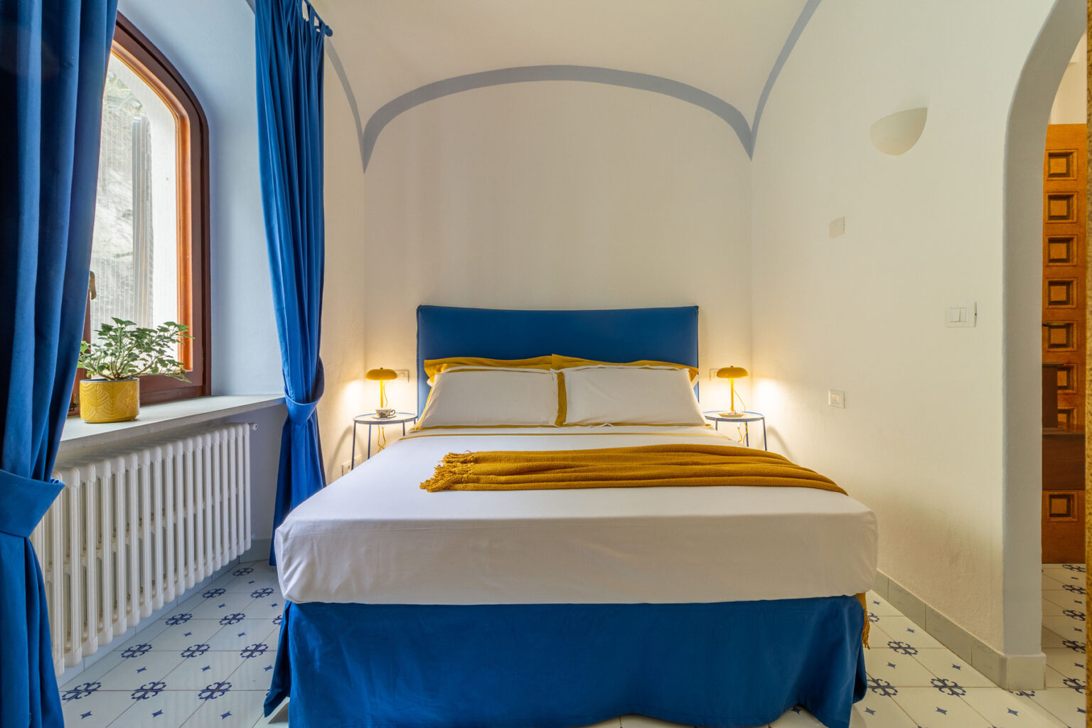 Cozy bedroom featuring a double bed with blue and white linens, warm yellow accents, and large windows with blue curtains, creating a bright and inviting atmosphere. | Only on SopranoVillas