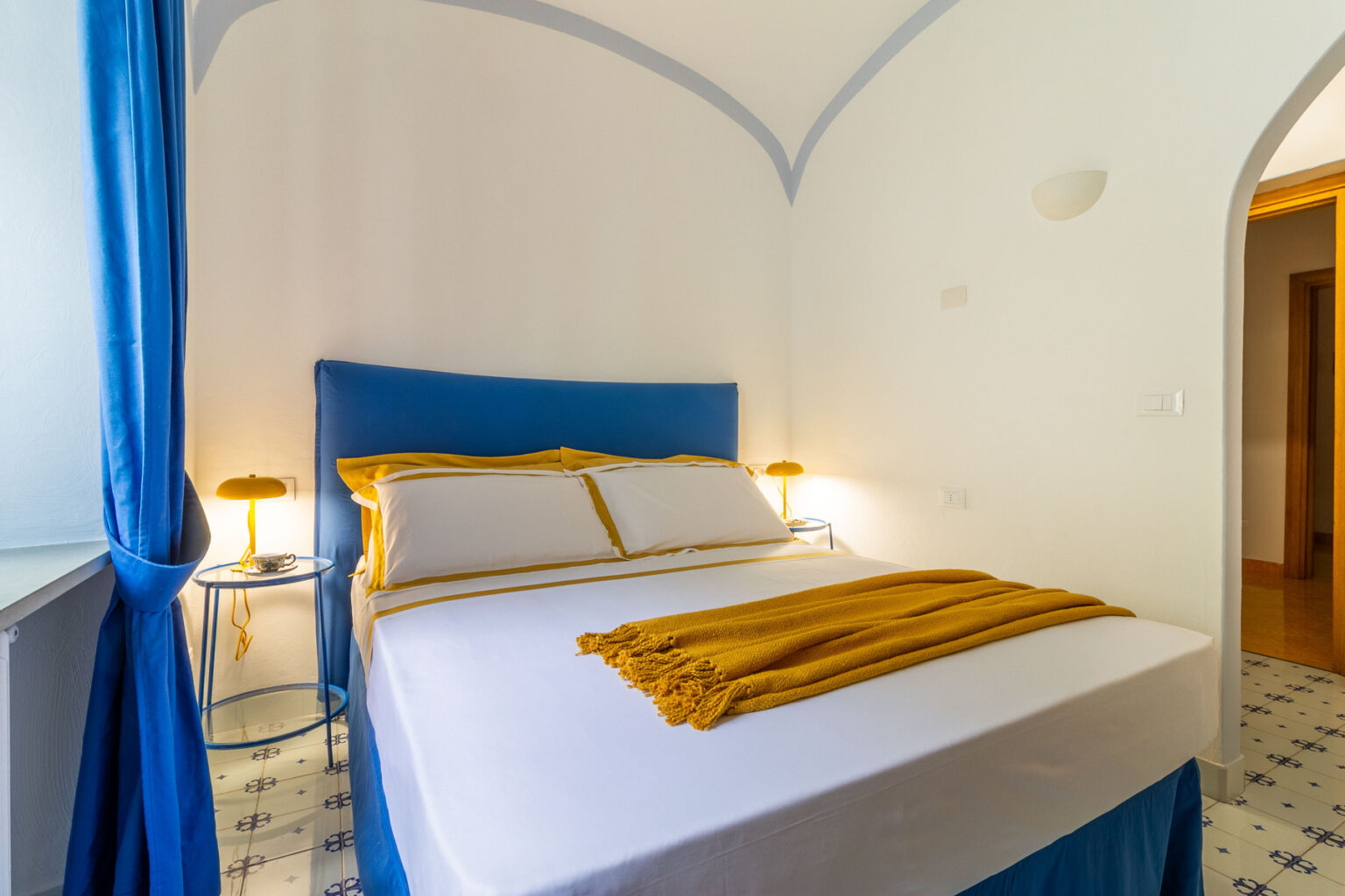 Cozy bedroom featuring a blue upholstered bed with yellow accents, white walls, and large windows with blue curtains, creating a bright and inviting atmosphere. Ideal for relaxation and comfort. | Only on SopranoVillas