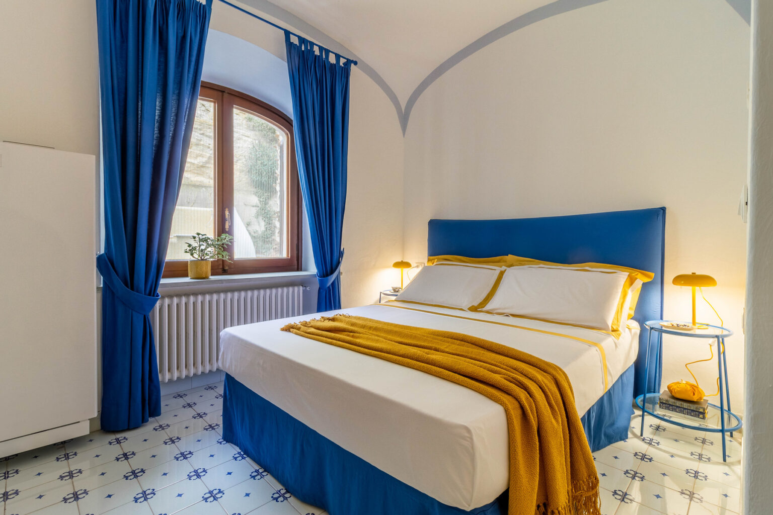 Cozy bedroom with a blue and white color scheme, featuring a comfortable bed with yellow accents, elegant curtains, and a small nightstand with a lamp, set against a bright window. Ideal for relaxation and tranquility. | Only on SopranoVillas