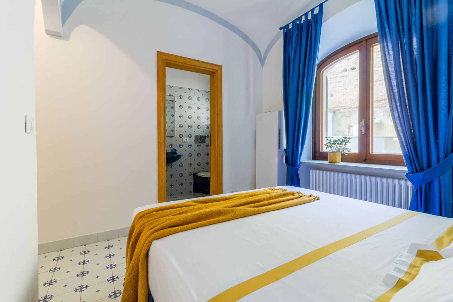 Cozy bedroom featuring a double bed with a yellow throw, blue curtains, and a bright window. The room has a modern design with tiled flooring and an en-suite bathroom visible through an open door, creating a welcoming atmosphere for relaxation. | Only on SopranoVillas