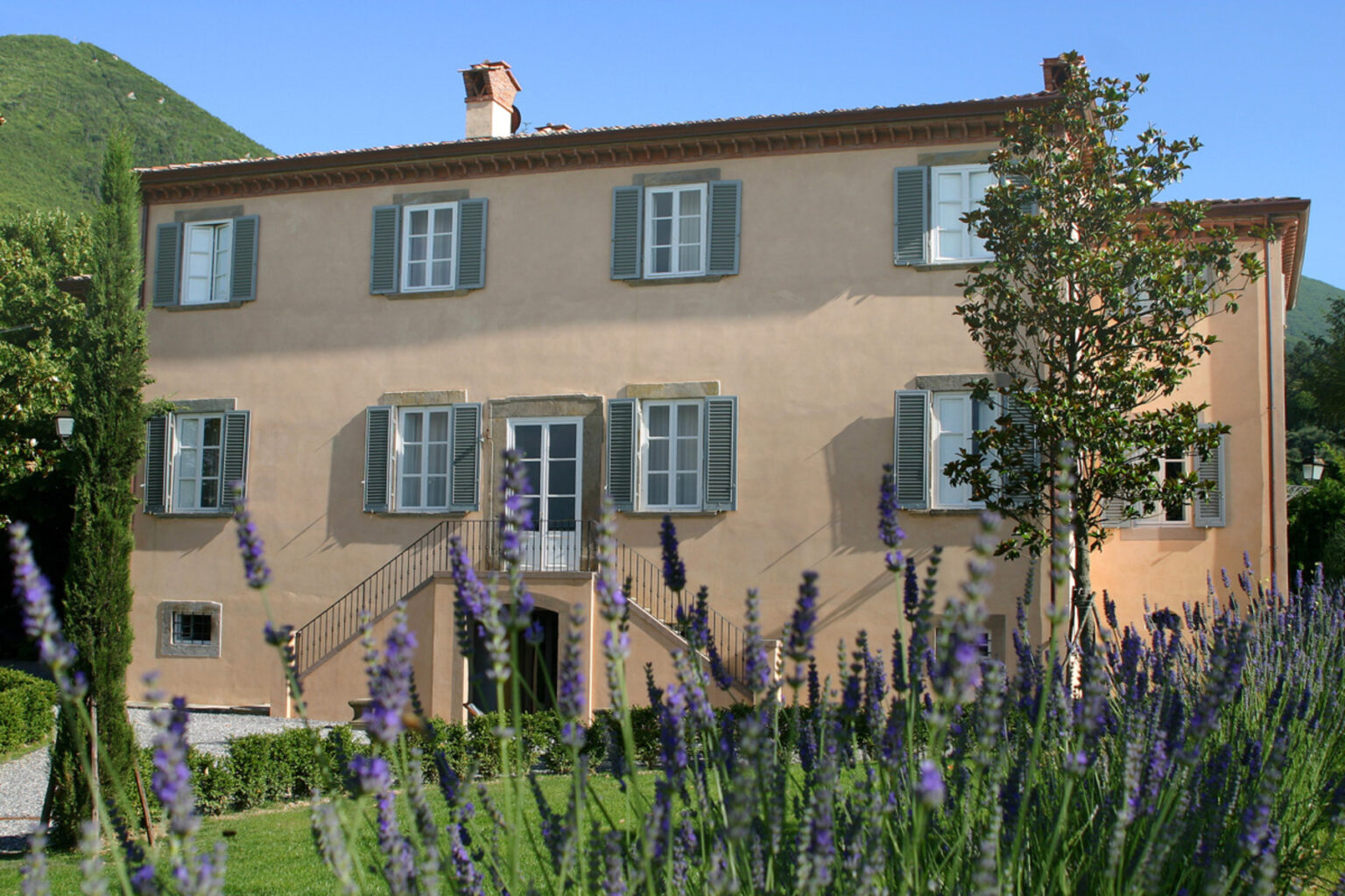 A charming two-story house with green shutters, surrounded by blooming lavender plants and lush greenery, set against a hillside backdrop. | Only on SopranoVillas