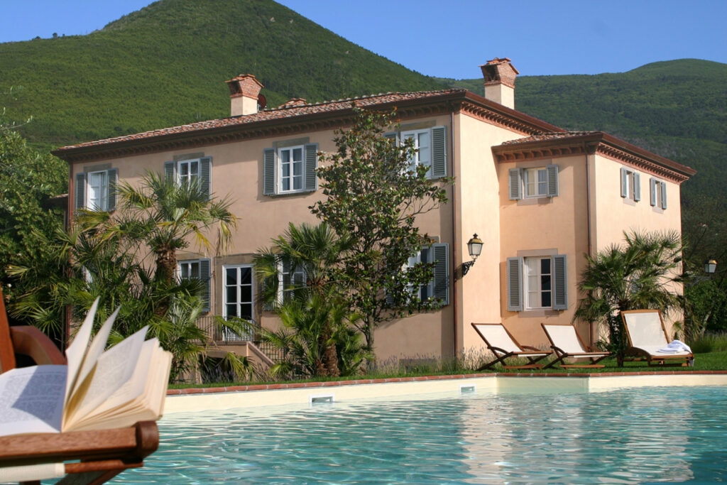 A beautiful villa surrounded by lush greenery and mountains, featuring a swimming pool in the foreground, lounge chairs, and an open book resting on a wooden deck chair, ideal for a relaxing getaway. | Only on SopranoVillas