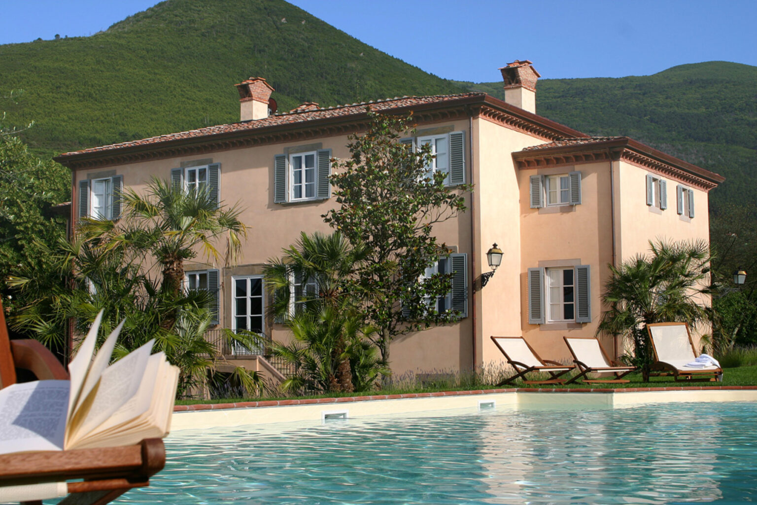A beautiful villa surrounded by lush greenery and mountains, featuring a swimming pool in the foreground, lounge chairs, and an open book resting on a wooden deck chair, ideal for a relaxing getaway. | Only on SopranoVillas