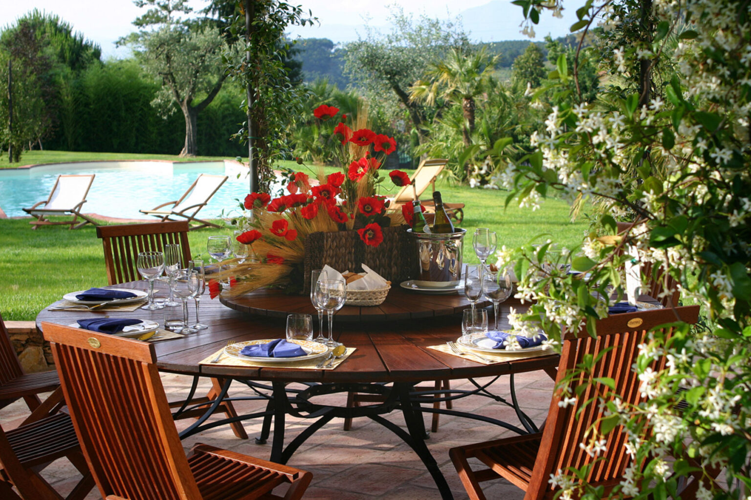 Outdoor dining setup featuring a round wooden table adorned with elegant dinnerware and a vibrant floral centerpiece. In the background, a serene swimming pool and lush greenery create a tranquil atmosphere, perfect for alfresco meals. | Only on SopranoVillas