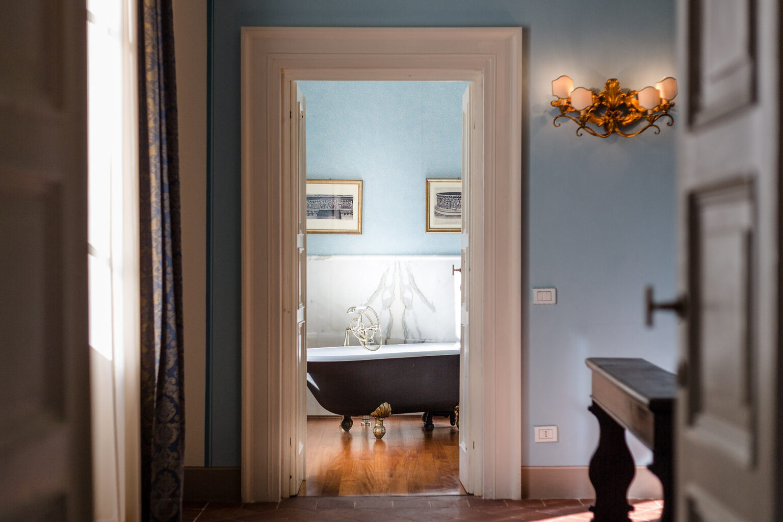 View of a stylish bathroom through an open doorway, featuring a vintage bathtub, elegant wall sconces, and soft blue walls, creating a serene atmosphere. | Only on SopranoVillas