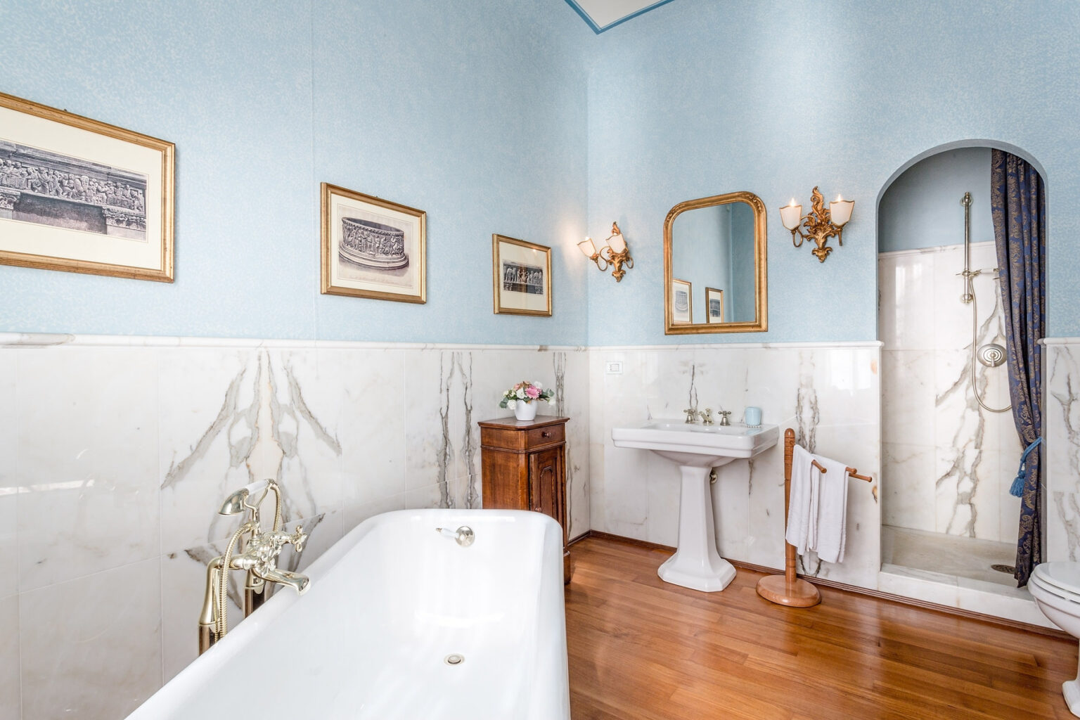 Elegant bathroom featuring a freestanding bathtub, vintage decor, and a pedestal sink. The walls are painted light blue with framed artwork, complemented by wooden flooring and stylish lighting fixtures. A shower area is visible in the corner, adding functionality to this charming space. | Only on SopranoVillas